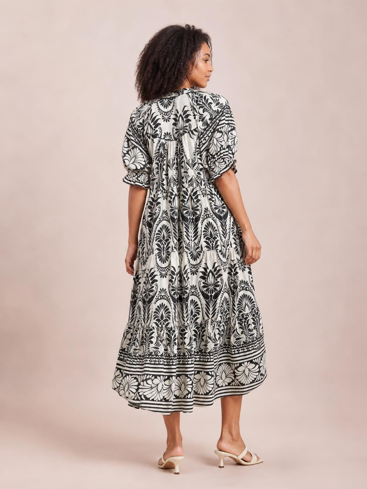 Model looks over her shoulder in a black-and-white two-tone border print midi dress with gathered tiers, elbow-length sleeves, and a flowing A-line silhouette.