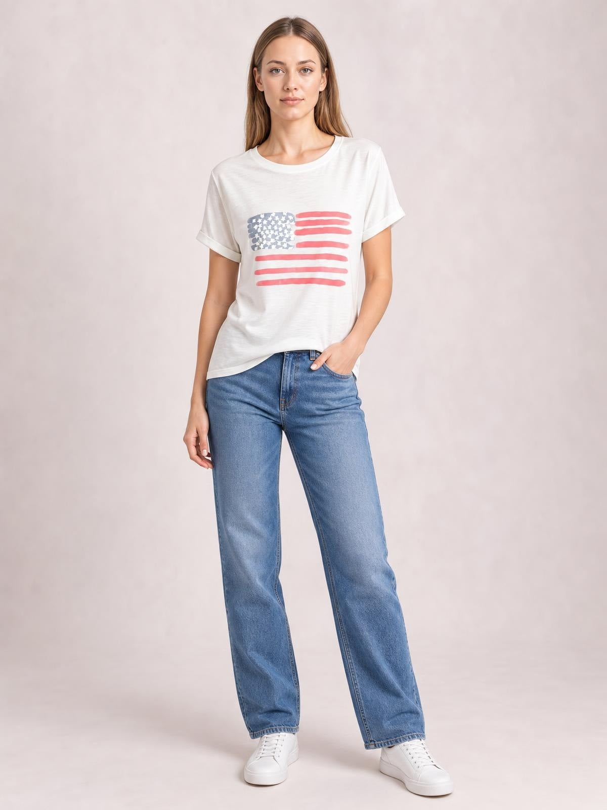 A model wears a white US flag round neck short sleeve T-shirt with rolled sleeve hems and a relaxed silhouette, standing with one hand in her jeans pocket.
