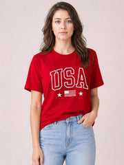 A model stands in a red USA American Flag crewneck T-shirt with short sleeves, a relaxed silhouette, graphic lettering, stars, and one hand in her jeans pocket.
