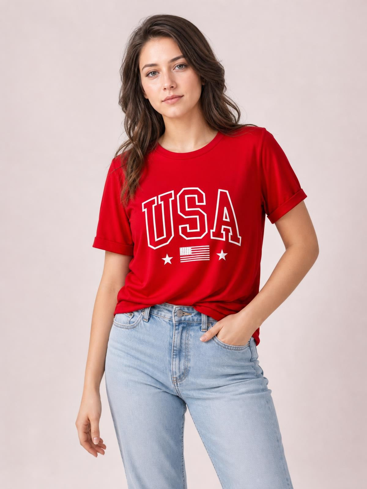 A model poses in a red USA American Flag crewneck T-shirt with rolled short sleeves, white graphic lettering, and a relaxed straight silhouette.
