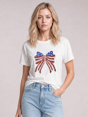 Model wears a white crew neck T-shirt with short sleeves, a relaxed silhouette, and a sparkling sequin USA flag bow knot patch, posing with one hand in her pocket.