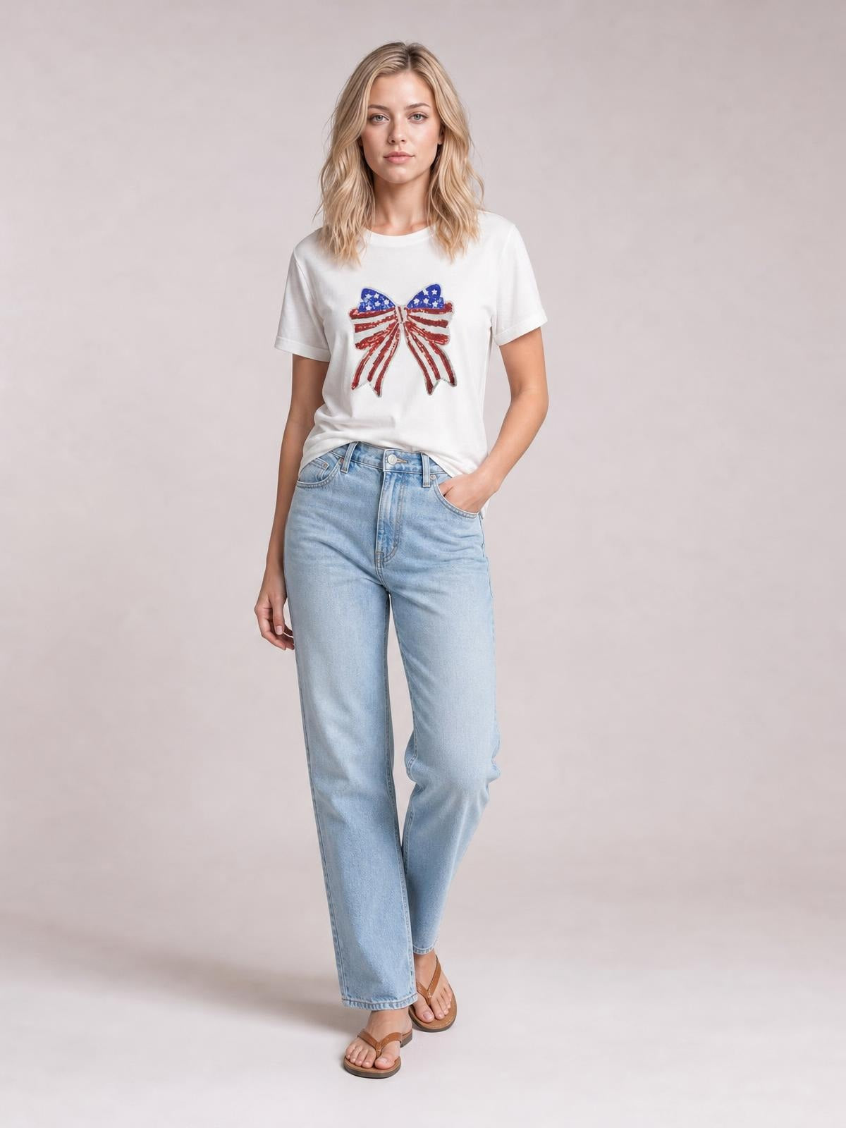 A model stands in a white crew neck T-shirt with short sleeves, a sparkling sequin USA flag bow knot patch, and a relaxed silhouette.