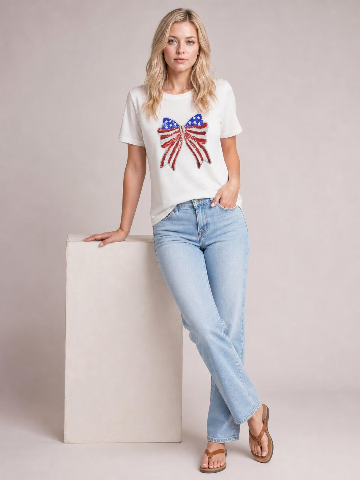 A model wears a white crew neck T-shirt with a sparkling sequin USA flag bow knot patch, short sleeves, and a relaxed silhouette while posing casually.