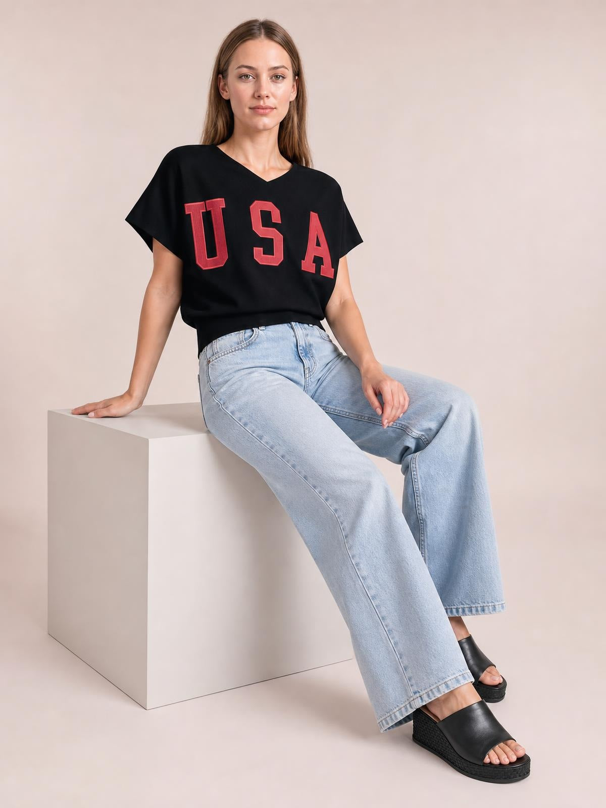 A seated model wears a black USA notched short-sleeve T-shirt with bold red lettering, a relaxed silhouette, and a V-shaped neckline.