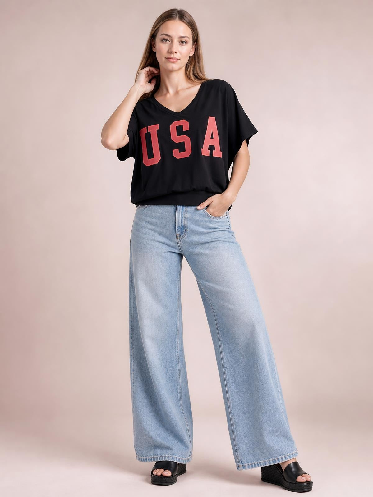 Model wears a black USA notched short-sleeve T-shirt with bold red lettering, a relaxed silhouette, and one hand resting near her neck.