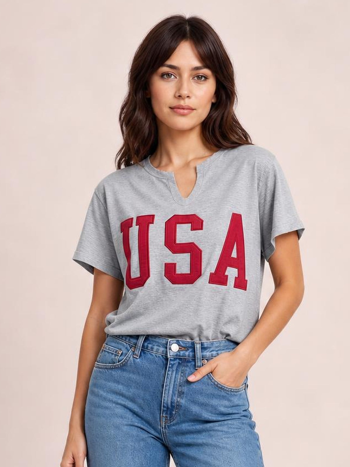 Model wearing a gray USA notched short-sleeve T-shirt with a relaxed silhouette, red varsity lettering, and a split neckline, posed with one hand in her jeans pocket.