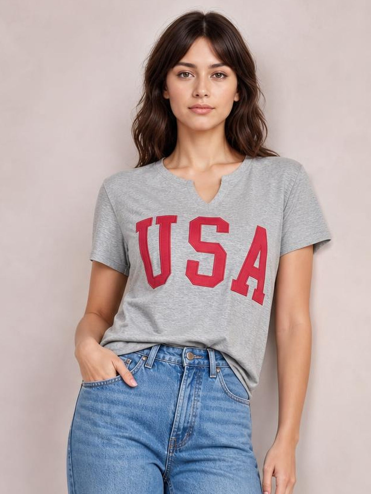 Model wears a gray USA notched short-sleeve T-shirt with a red varsity-style graphic, relaxed silhouette, and one hand resting in her jeans pocket.