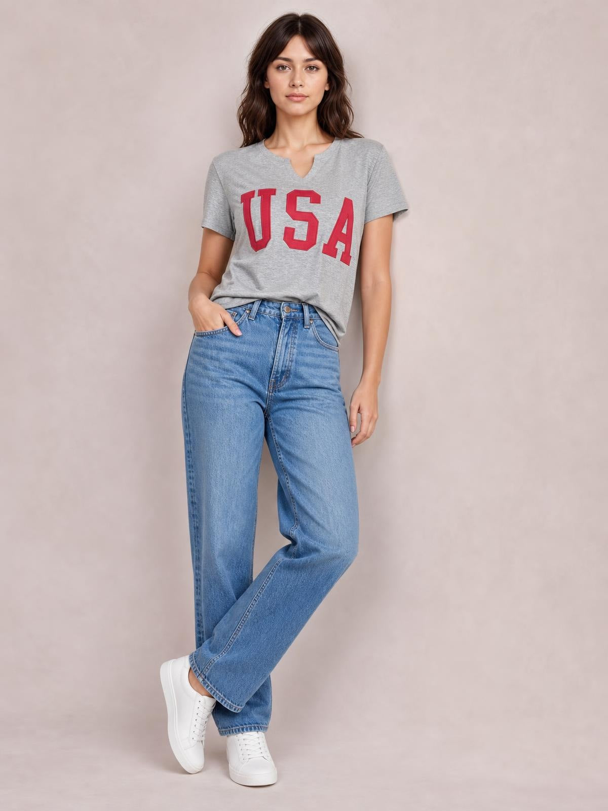 A model wears a gray USA notched short sleeve T-shirt with bold red lettering and a relaxed silhouette, standing with one hand in her jeans pocket.