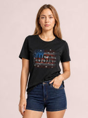 Model wears a black USA rhinestone round-neck T-shirt with short sleeves and a relaxed silhouette, paired with denim shorts and one hand in a pocket.