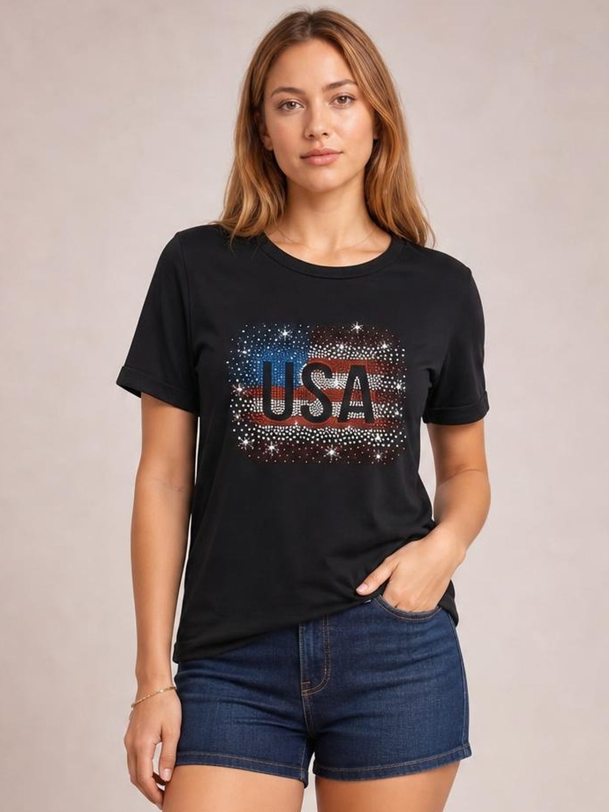 Model wearing a black USA rhinestone T-shirt with a round neck, short sleeves, and relaxed silhouette, posing with one hand in her denim shorts pocket.