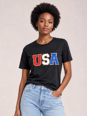 Model poses with one hand in her pocket wearing a black USA round neck short sleeve T-shirt with a relaxed silhouette and bold red, white, and blue lettering.
