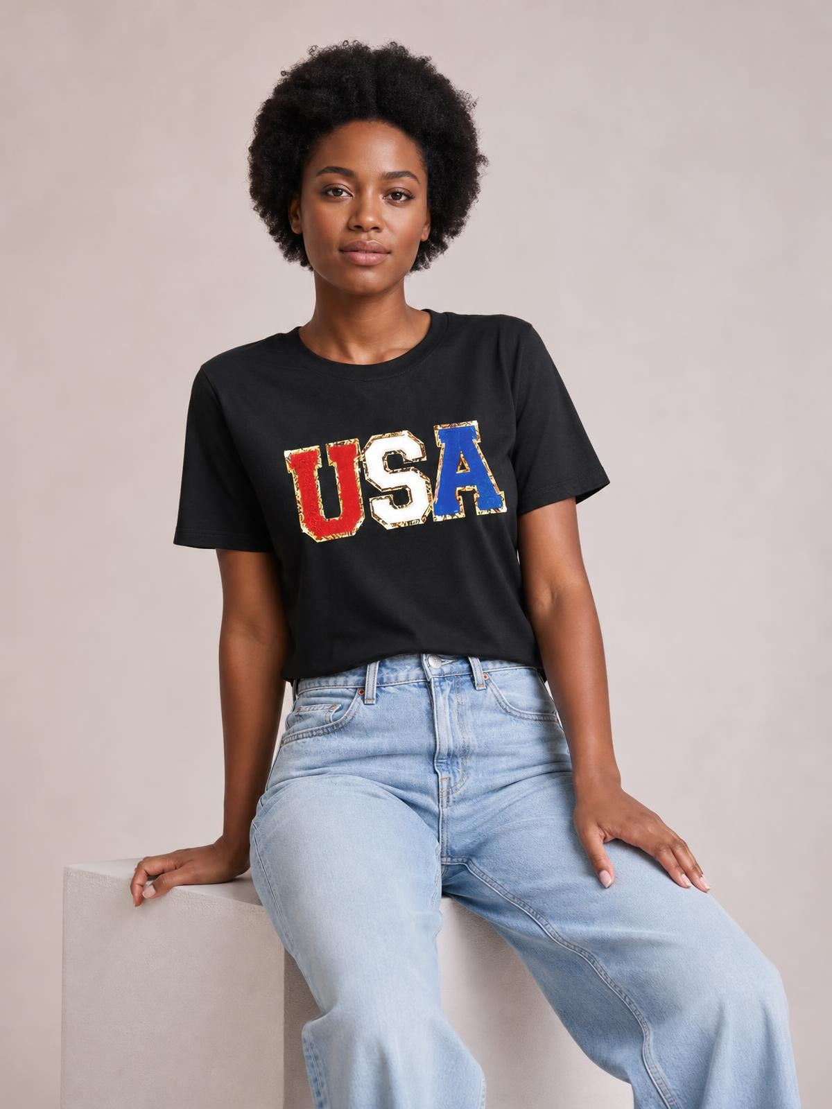 Model seated in a relaxed pose wearing a black USA round neck short sleeve T-shirt with a bold multicolor graphic and straight silhouette.