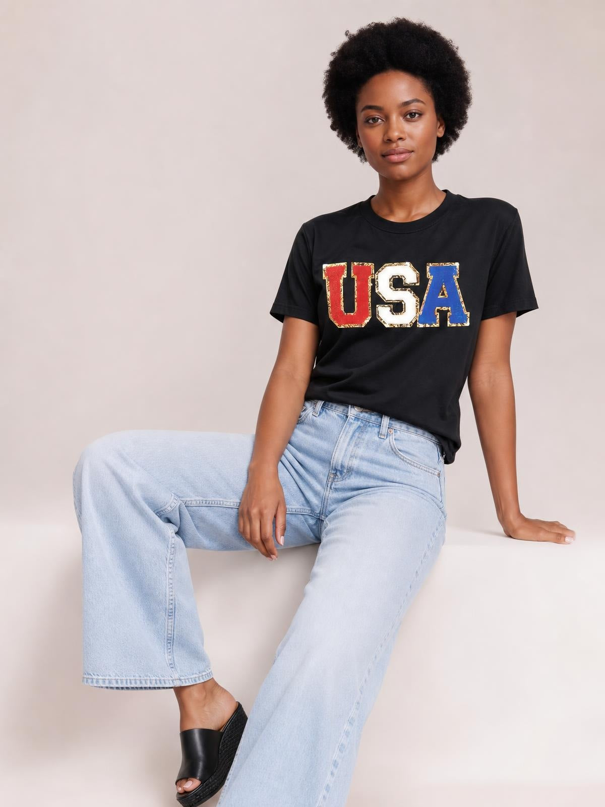 A seated model wears a black USA round neck short sleeve T-shirt with a bold multicolor graphic, relaxed silhouette, and ribbed crew neckline.
