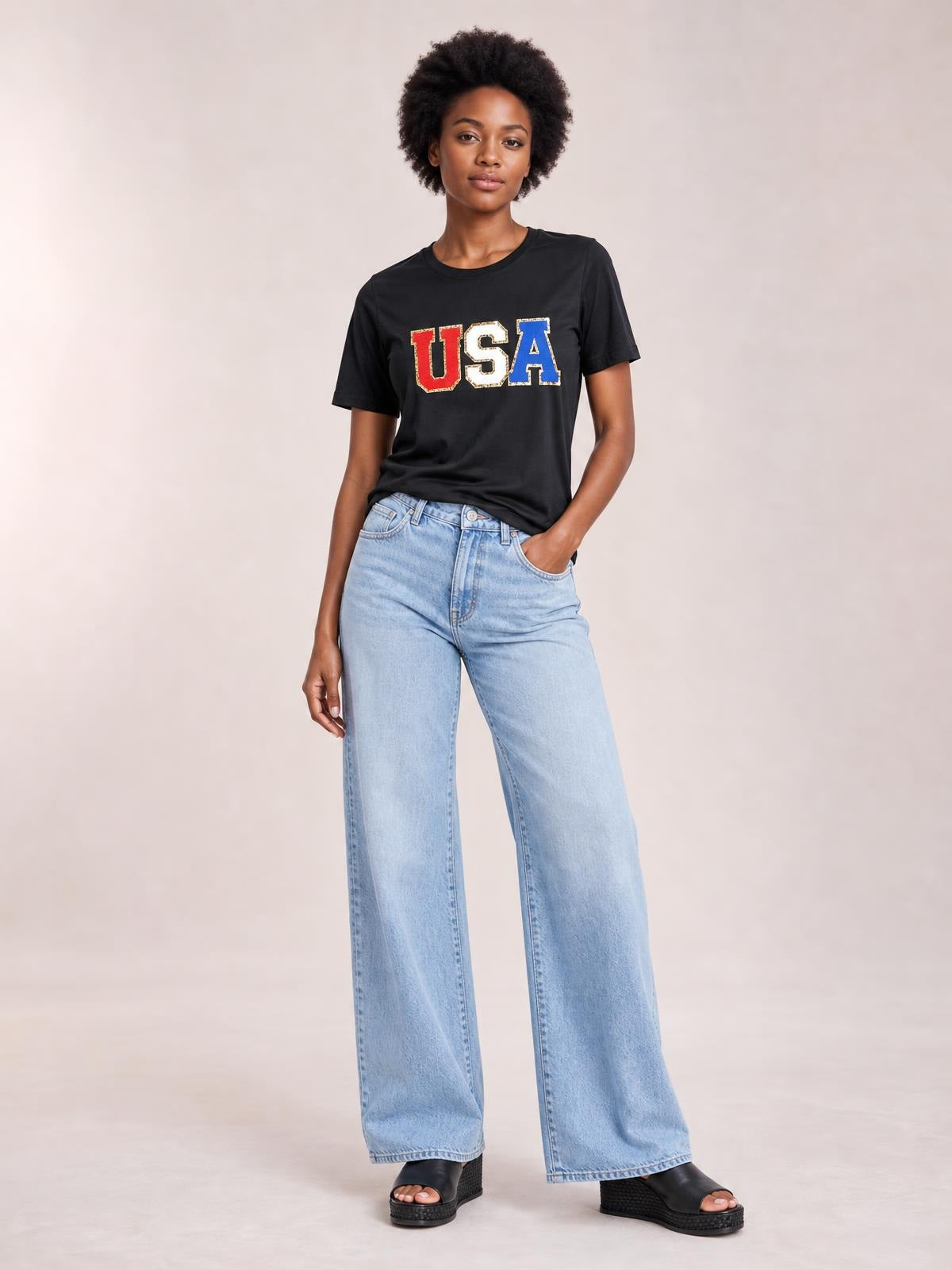 Model poses in a black USA round neck short sleeve T-shirt with a straight silhouette, bold multicolor USA lettering, and one hand tucked into her jeans pocket.