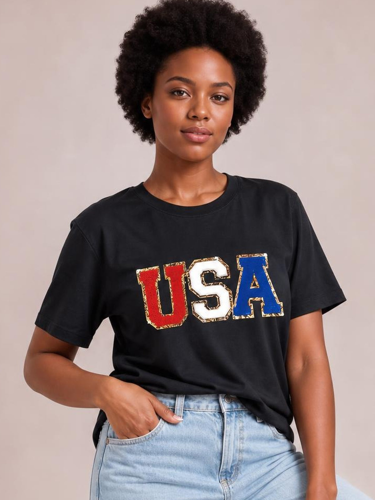 Model wearing a black USA round neck short-sleeve T-shirt with bold red, white, and blue lettering, shown in a relaxed pose with one hand in her jeans pocket.