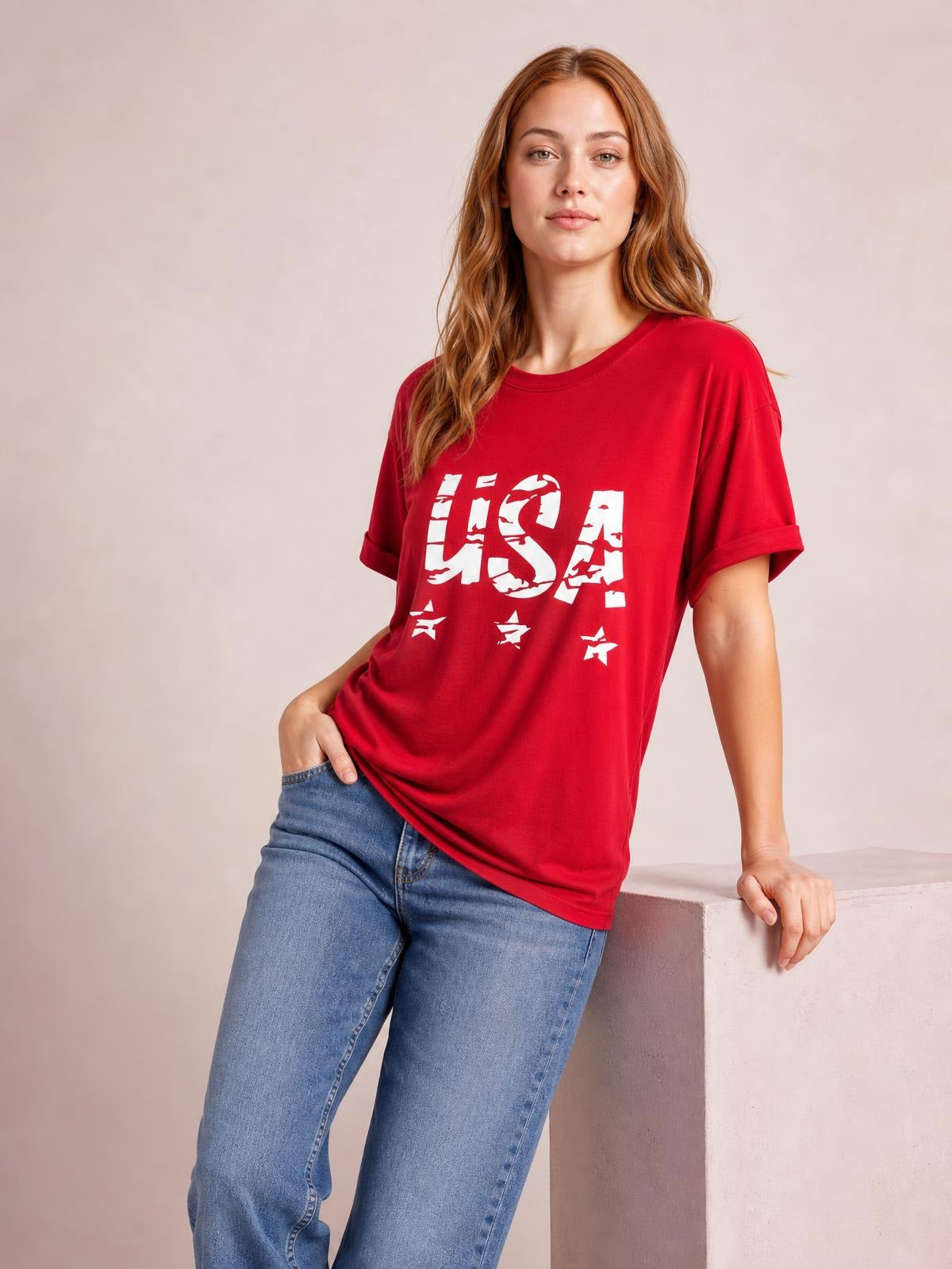 A model leans against a ledge wearing a red USA round-neck short-sleeve T-shirt with rolled cuffs, a relaxed classic fit, and bold white star graphics.