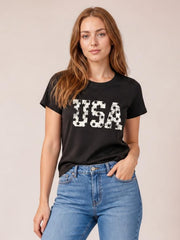 Model wearing a black USA round neck short sleeve T-shirt with a bold white star-print graphic and relaxed silhouette, standing with one hand in her jeans pocket.