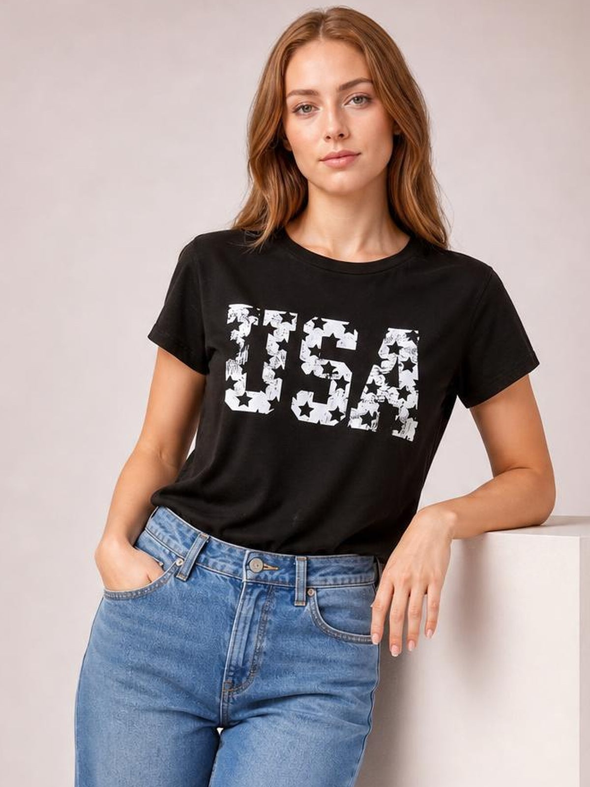 Model wearing a black USA round neck short sleeve T-shirt with a relaxed silhouette and distressed white star graphic, posed with one hand in her jeans pocket.