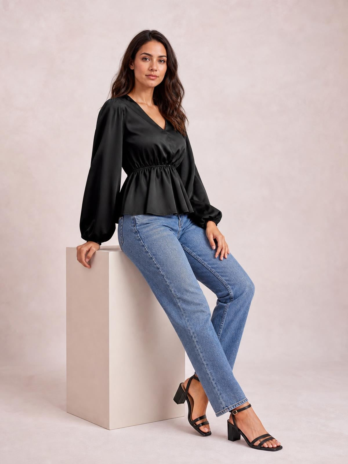 Model seated in a black V-neck peplum blouse with voluminous balloon sleeves, gathered waist, and flared hem, paired with jeans.
