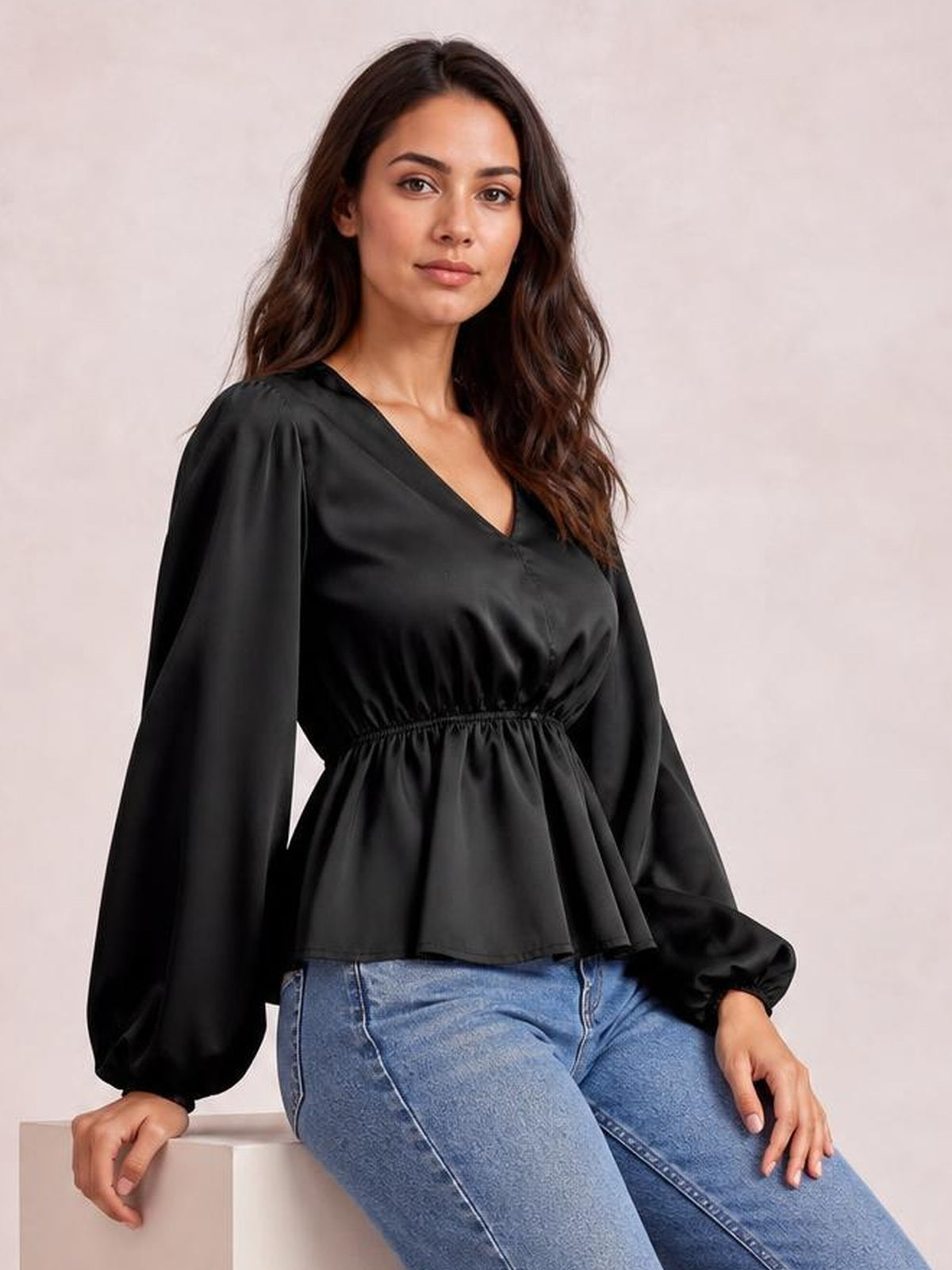 Model seated in a black V-neck peplum blouse with voluminous balloon sleeves, gathered waist, and flared hem, paired with blue jeans.
