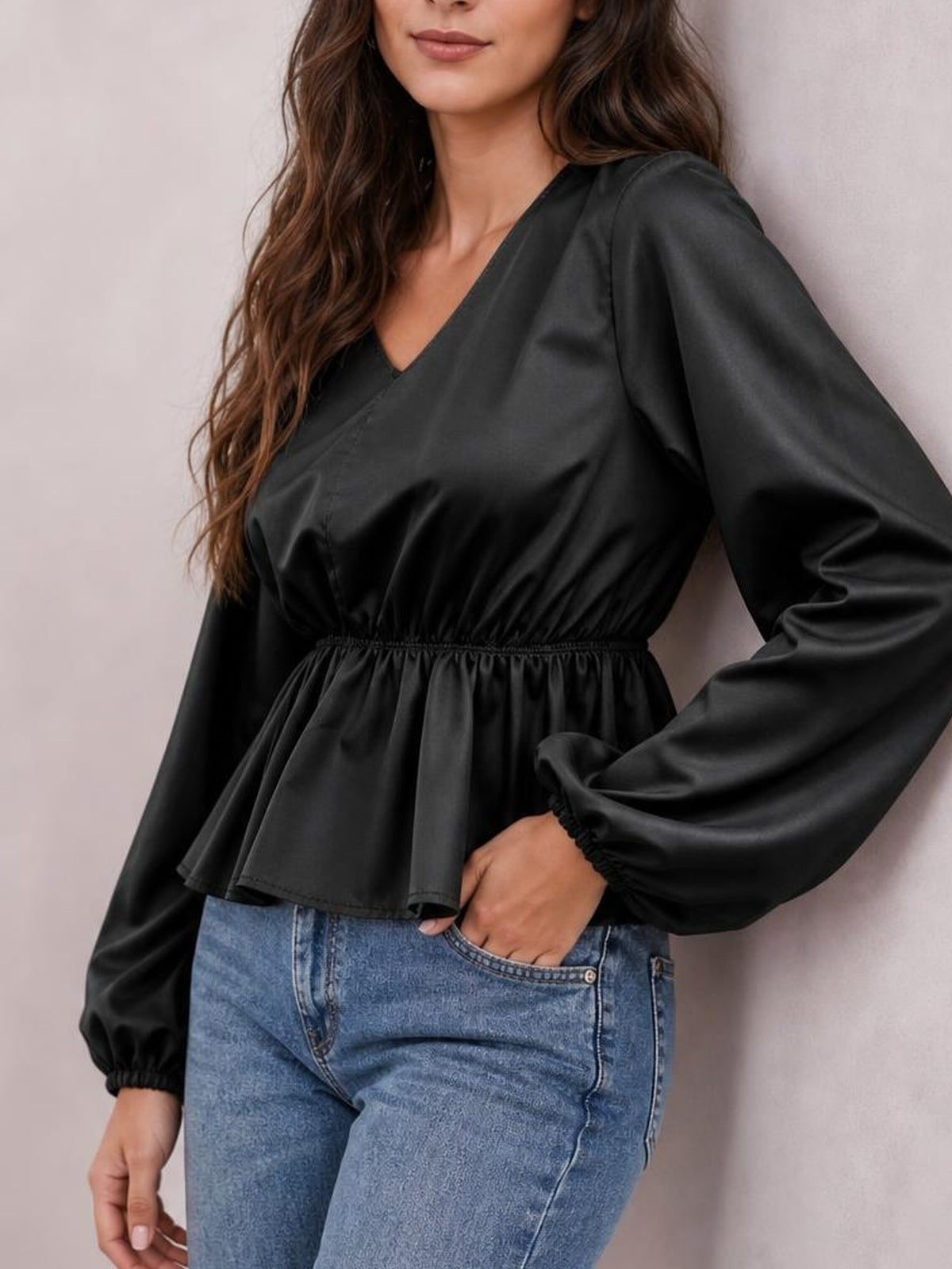 A model poses with one hand in her jeans pocket wearing a black V-neck peplum blouse with voluminous balloon sleeves and elastic cuffs.