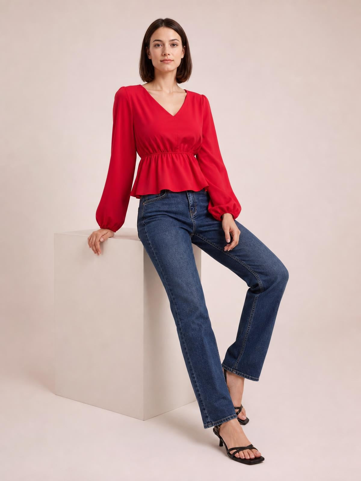 A model sits in a red V-neck peplum blouse with voluminous balloon sleeves, gathered waist, and flared hem, paired with blue jeans.