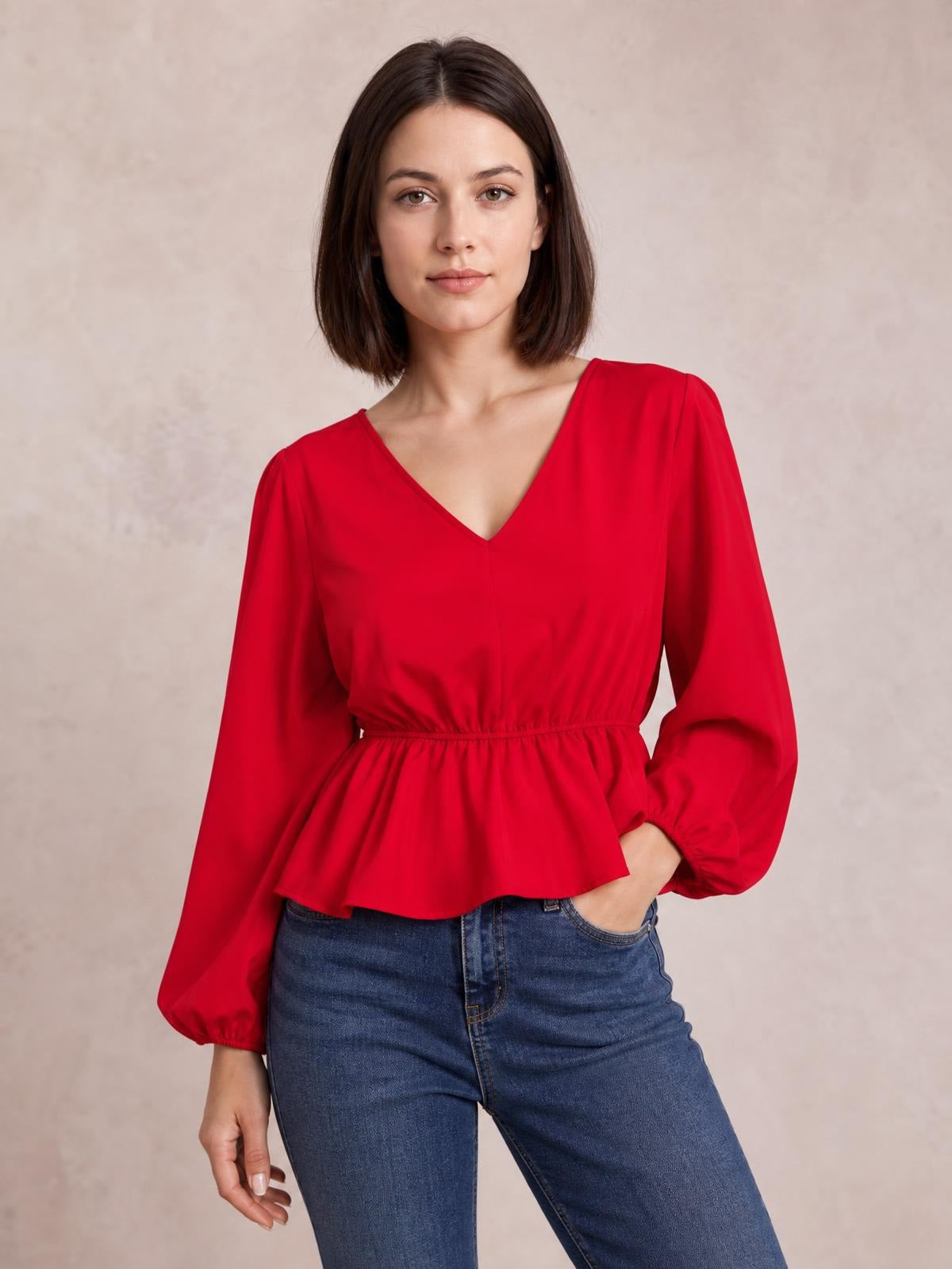 Model wears a red V-neck peplum blouse with gathered waist and voluminous balloon sleeves, paired with jeans in a relaxed standing pose.