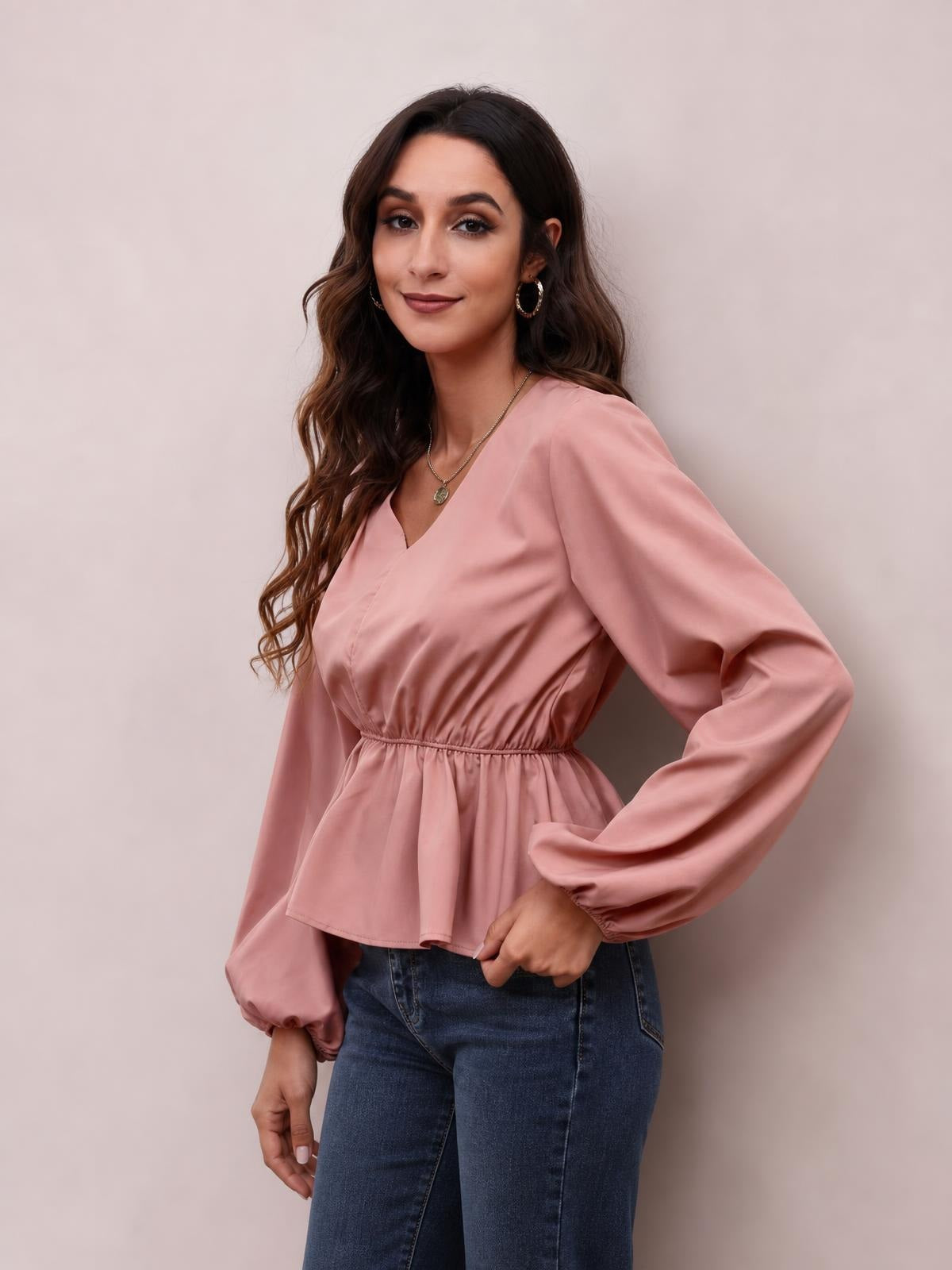 Model poses with one hand in her pocket wearing a pink V-neck peplum blouse with voluminous balloon sleeves, gathered waist, and flared hem.