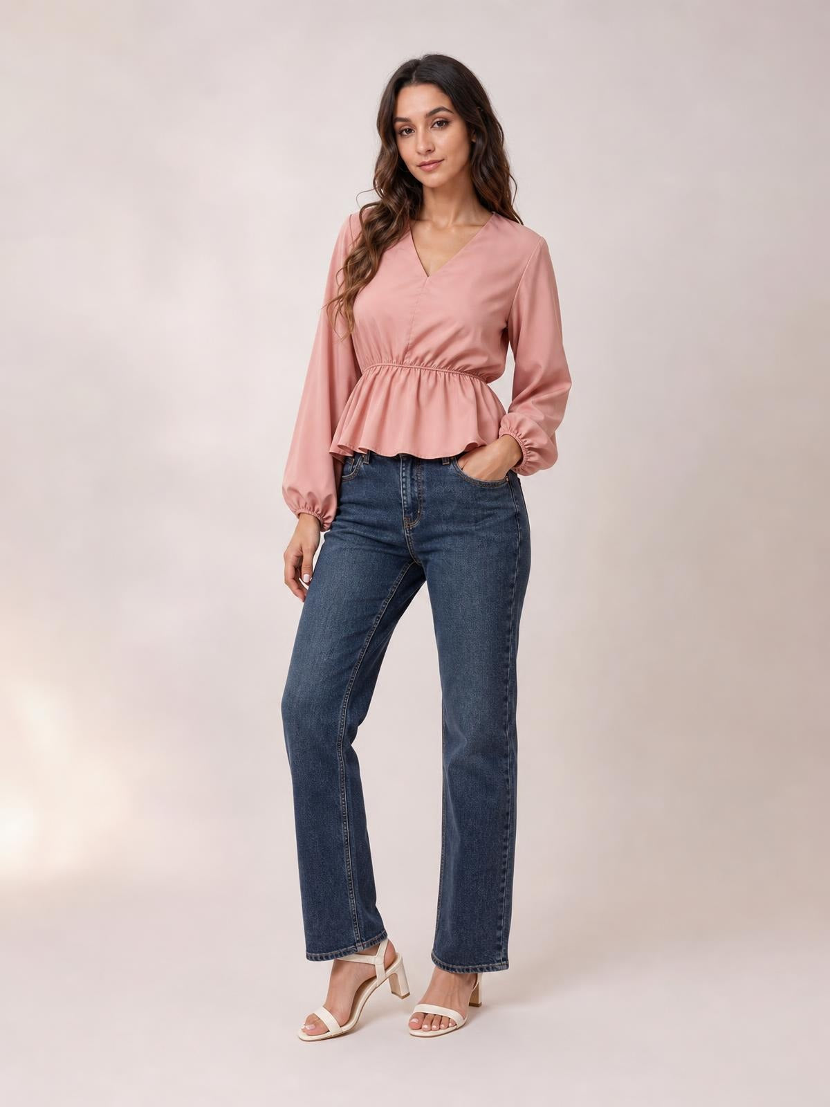 A model wears a pink V-neck peplum blouse with long balloon sleeves, a gathered waist, and elastic cuffs, posing with one hand in a jeans pocket.