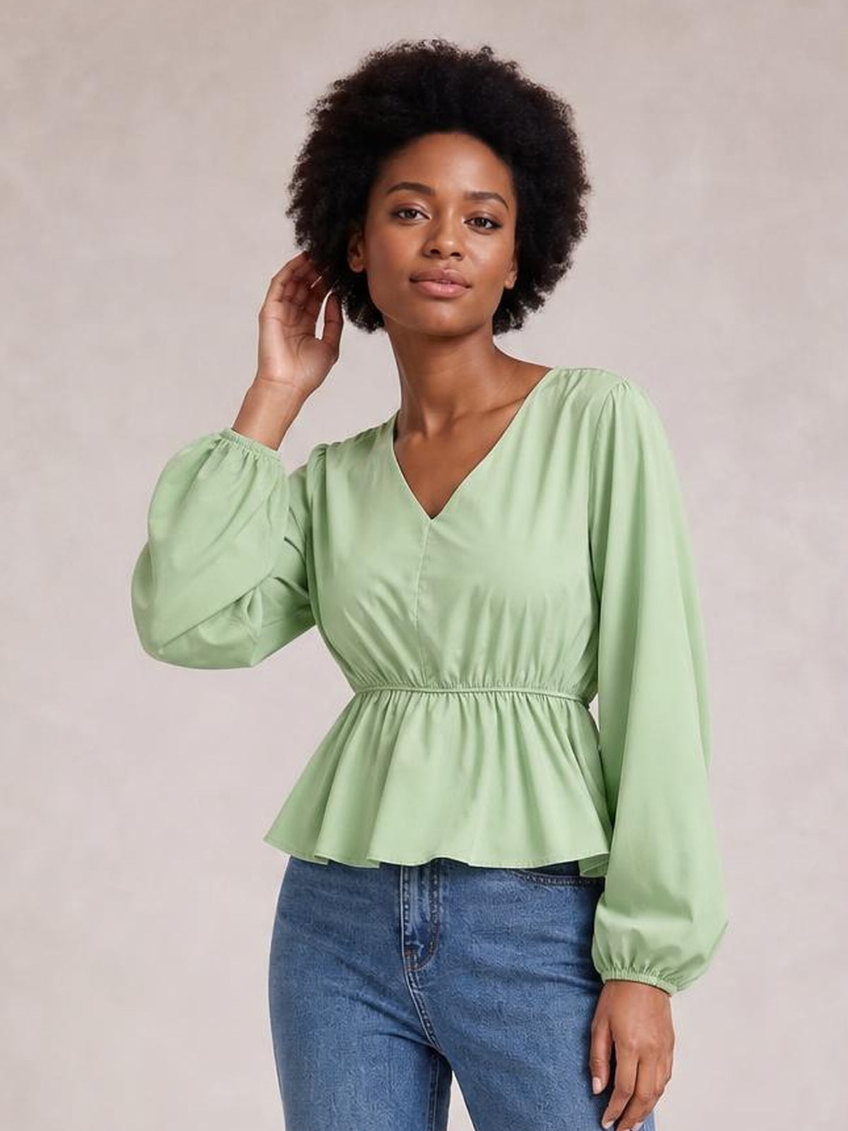 Model wearing a sage green V-neck peplum blouse with voluminous balloon sleeves, gathered cuffs, an elastic waist, and flared hem, posing with one hand in her hair.