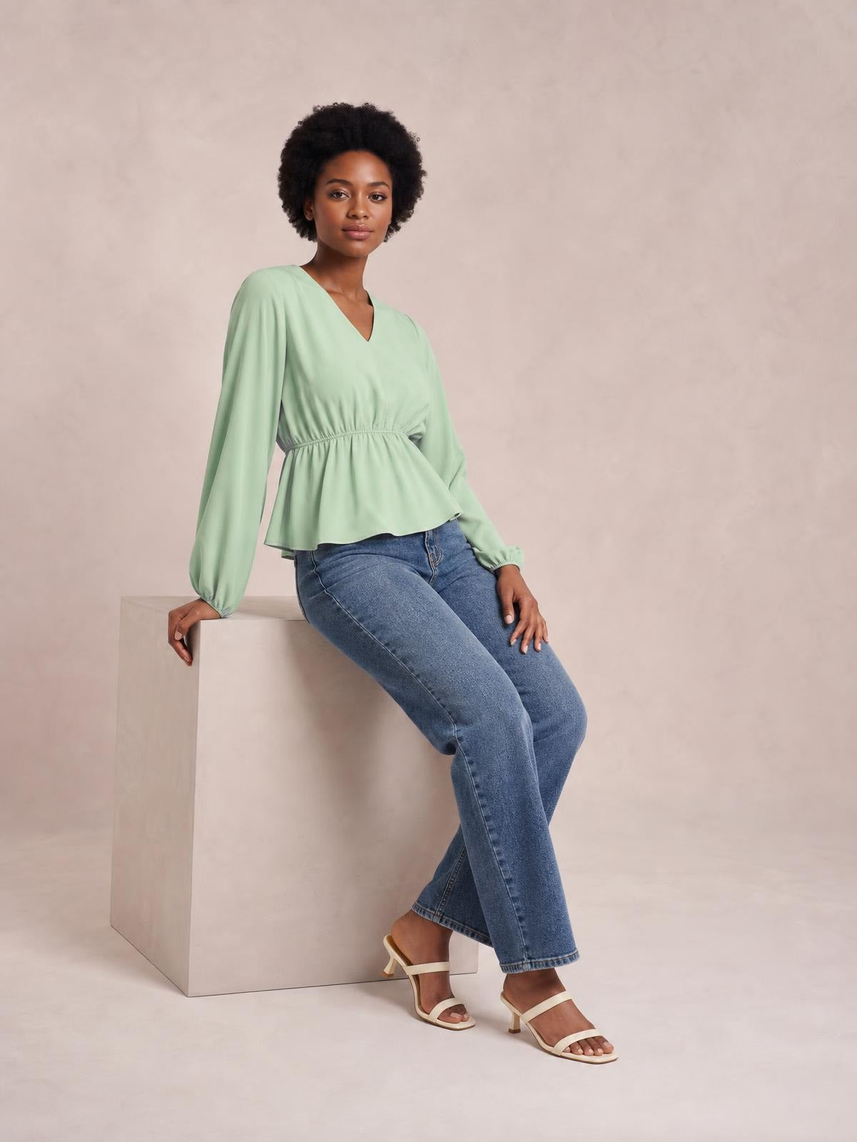 Model seated in a mint green V-neck peplum blouse with long balloon sleeves, gathered waist, and flared hem, paired with blue jeans.