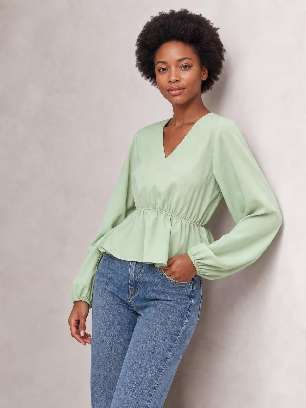 Model poses with one hand in her pocket wearing a light green V-neck peplum blouse with balloon sleeves, elastic cuffs, and a defined waist.