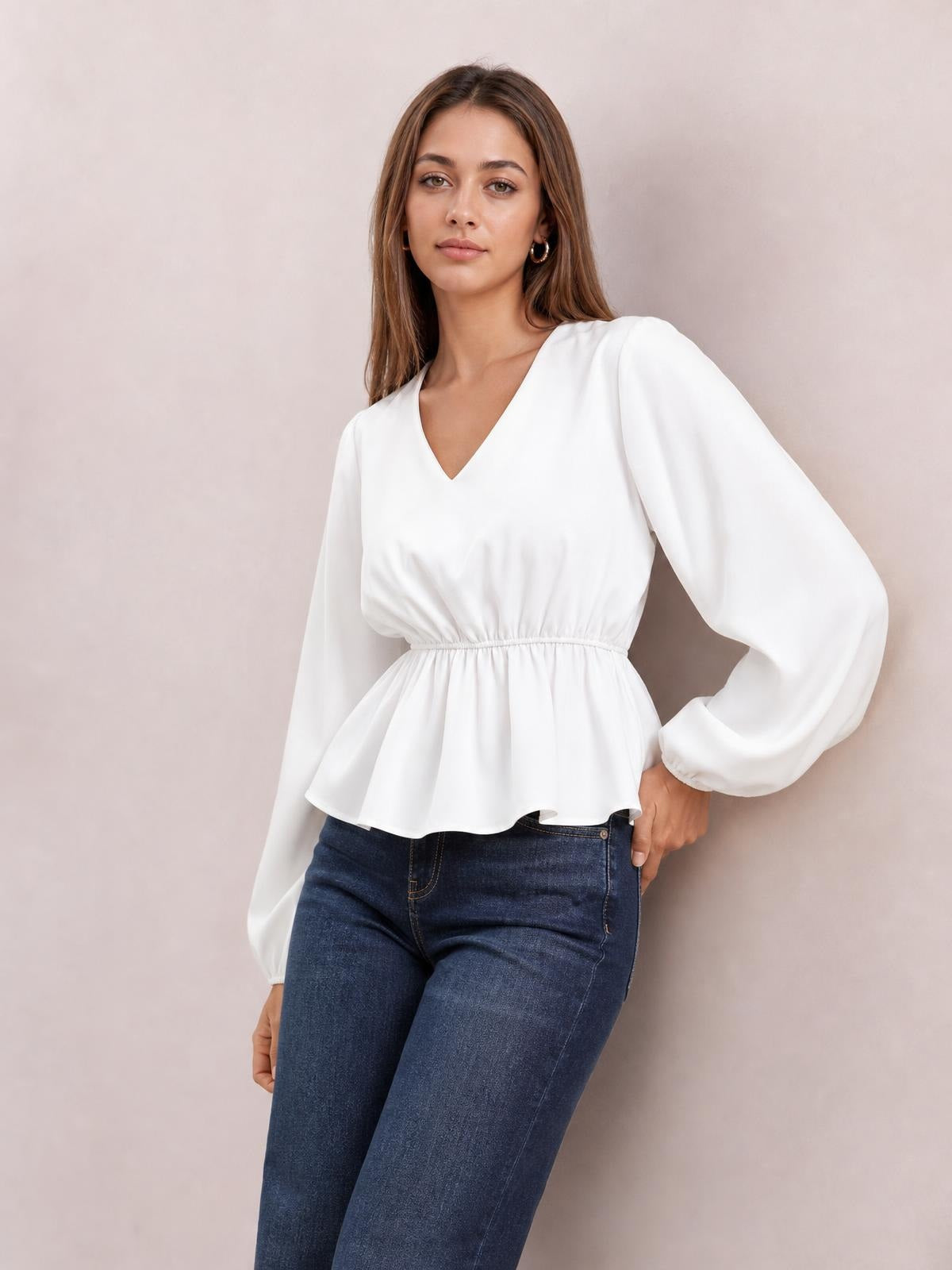 Model poses in a white V-neck peplum blouse with voluminous balloon sleeves, gathered waist, and flared hem, paired with dark jeans.