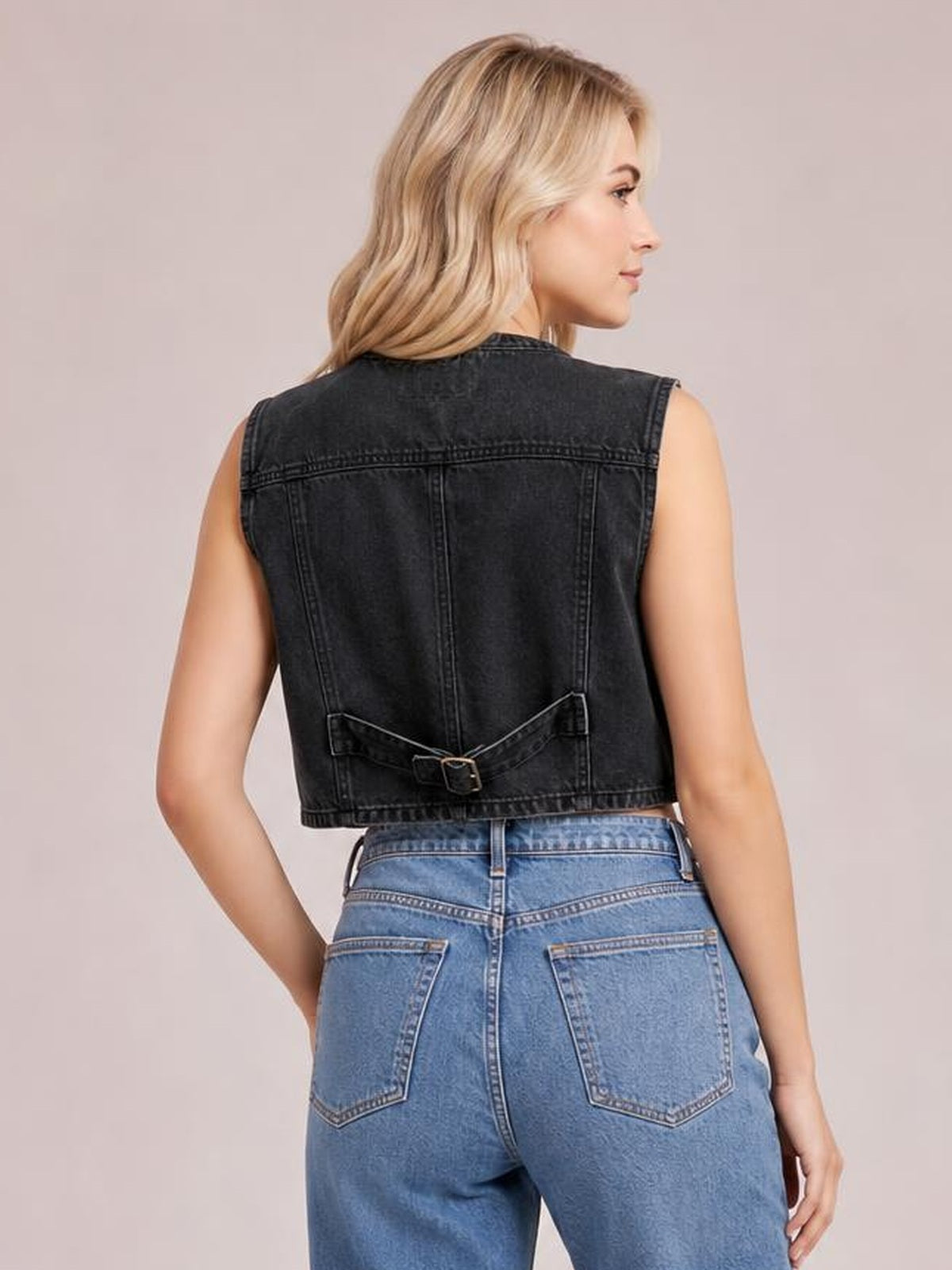 Model shown from the back in a black denim vest with a sleeveless cropped silhouette, seamed panel construction, and adjustable buckle detail at the waist.
