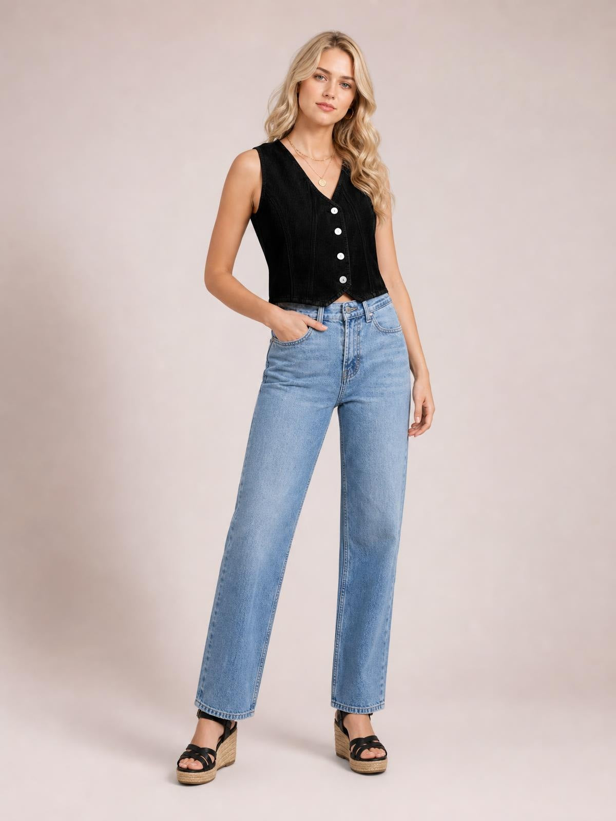 A standing model wears a black V-neck button-down vest with a cropped, sleeveless silhouette and visible front buttons, one hand in her jeans pocket.