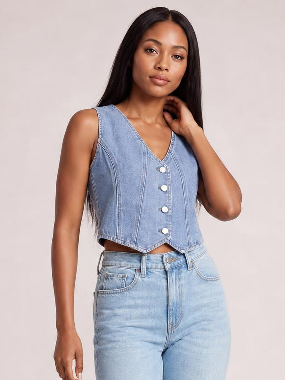 Model poses in a light blue V-neck button-down denim vest with sleeveless styling, tailored seam details, a fitted cropped silhouette, and pointed hem.