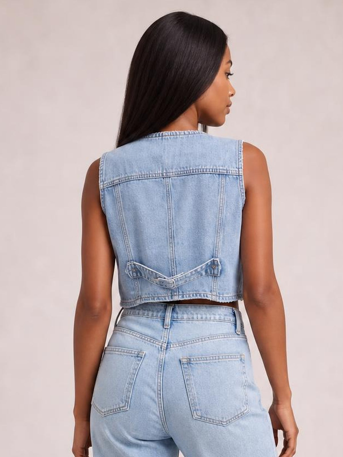 Model shown from the back wearing a light-wash denim vest with a cropped silhouette, sleeveless cut, contrast seams, and an adjustable back buckle.