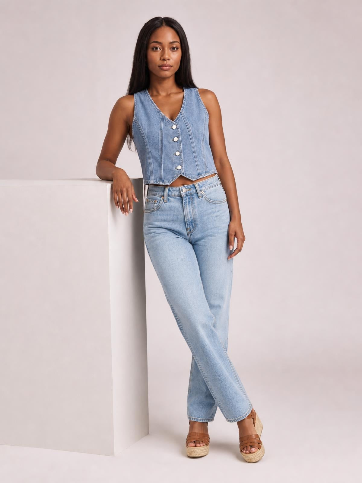 Model poses in a light blue denim vest with a V-neck, sleeveless cut, button-down front, shaped seams, and cropped hem.