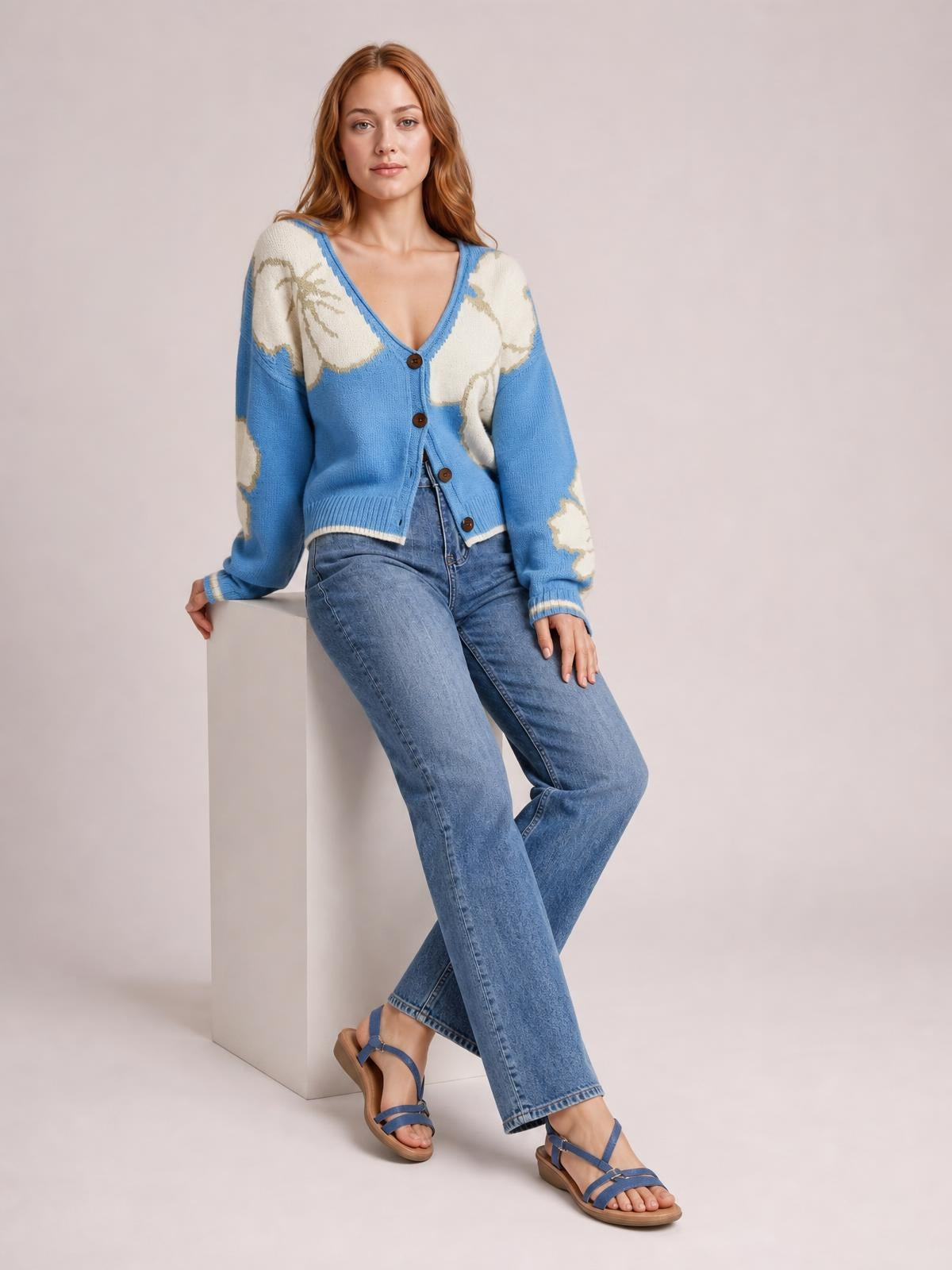 Model seated with crossed legs wears a blue V-neck button-down knit cardigan featuring cream floral panels, long sleeves, ribbed trim, and a relaxed silhouette.