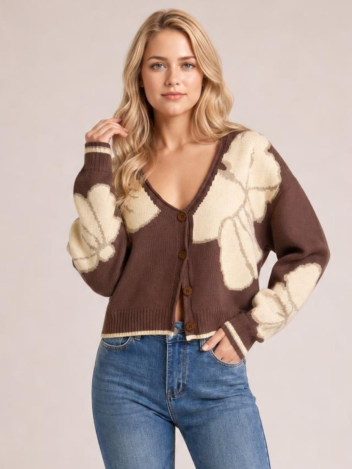 Model wearing a brown and cream knit cardigan with a V-neck, button-down front, contrast seam details, ribbed trim, and a cropped silhouette, posing with one hand in her jeans pocket.