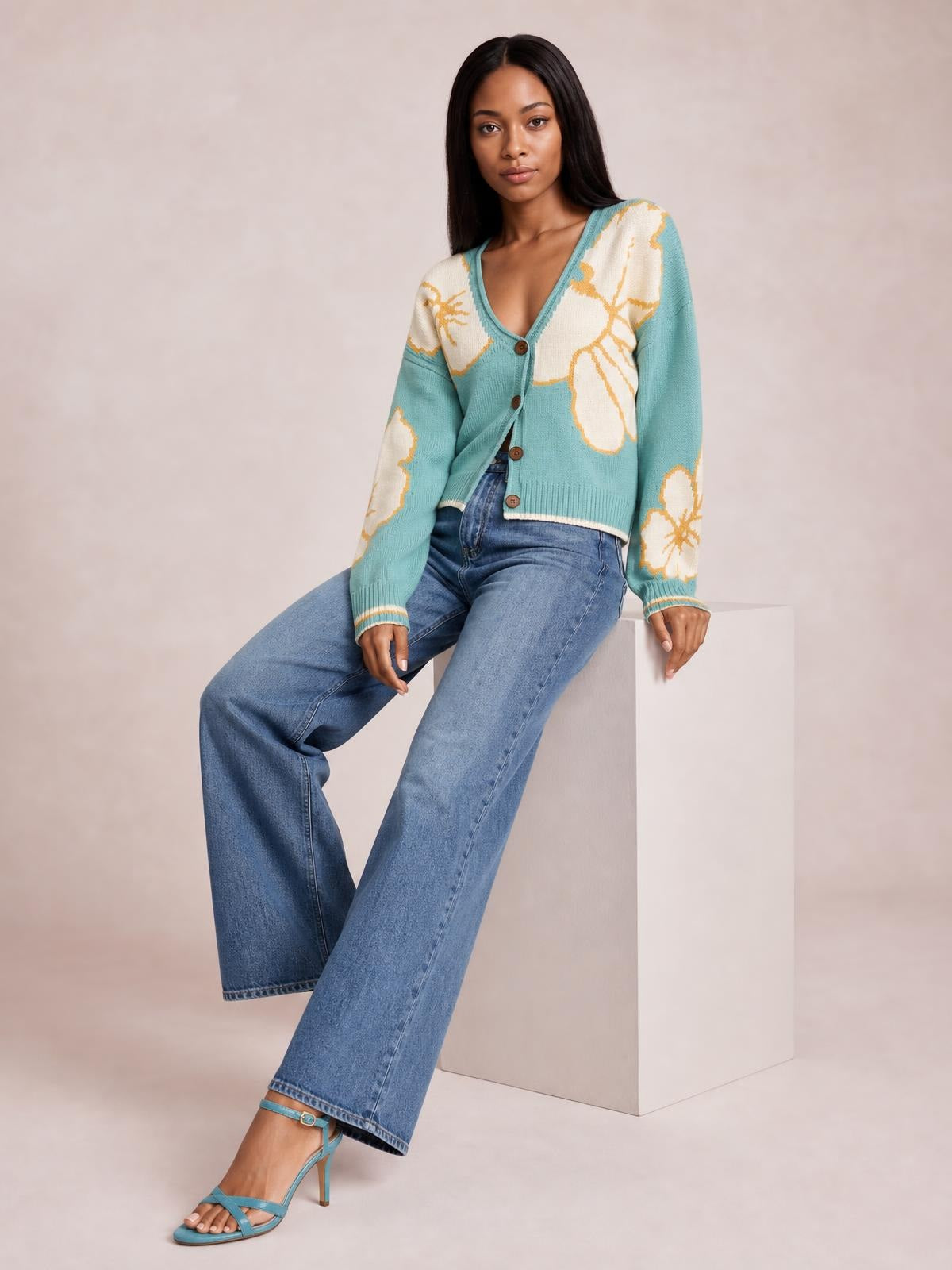 A seated model wears a turquoise V-neck button-down knit cardigan with cream floral motifs, ribbed cuffs and hem, and a cropped relaxed silhouette.