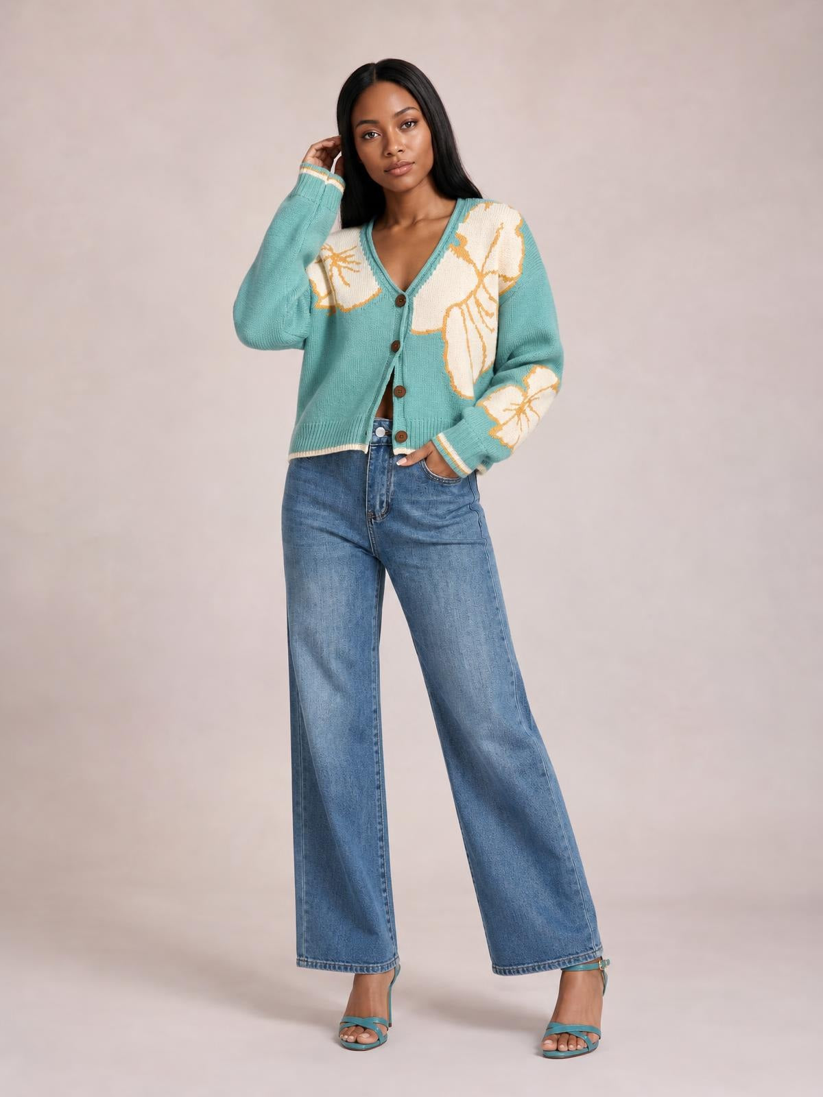 A model poses in a turquoise V-neck button-down knit cardigan with white and tan floral motifs, seam detail, ribbed trim, and a relaxed silhouette.