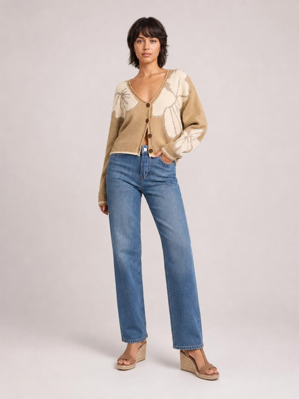 A model poses with one hand in her pocket wearing a tan and cream knit cardigan with a V-neck, button-down front, long sleeves, and ribbed trim.