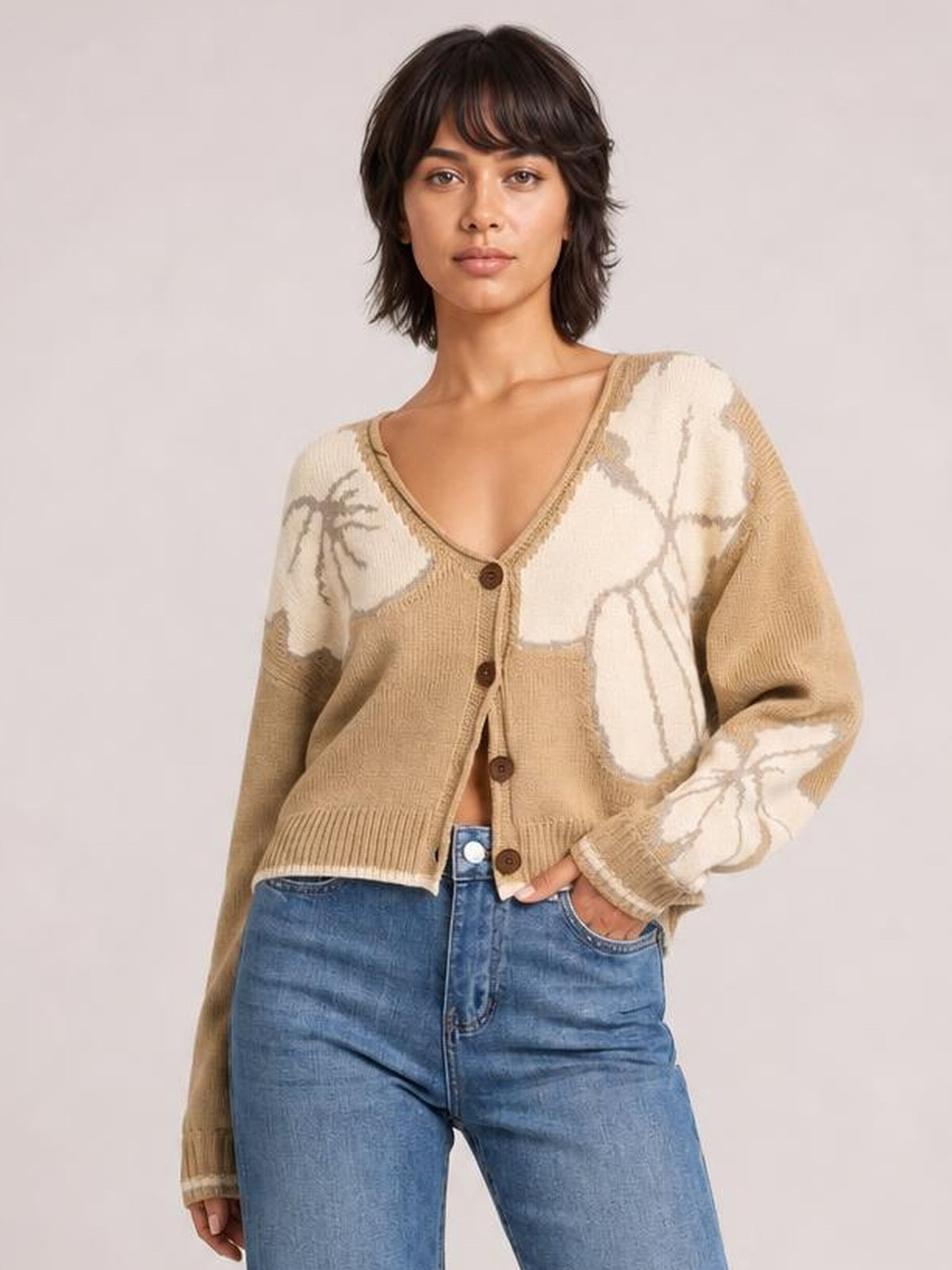 Model poses with one hand in her jeans pocket, wearing a beige V-neck button-down knit cardigan with brown buttons, seam detail, ribbed cuffs, and cream abstract patches.
