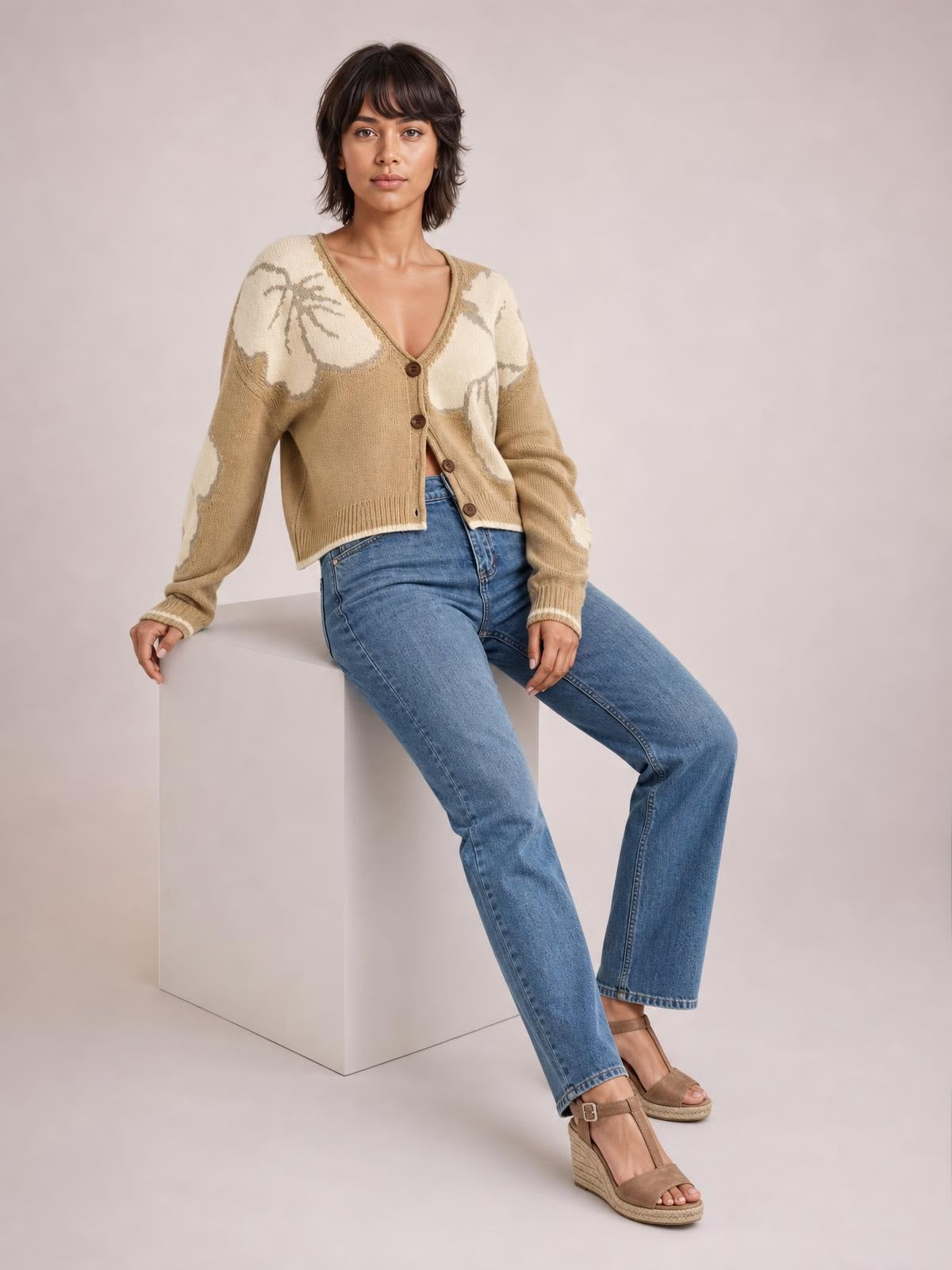 Model seated in a tan and cream knit cardigan with a V-neck, button-down front, seam detail, long sleeves, and cropped silhouette.