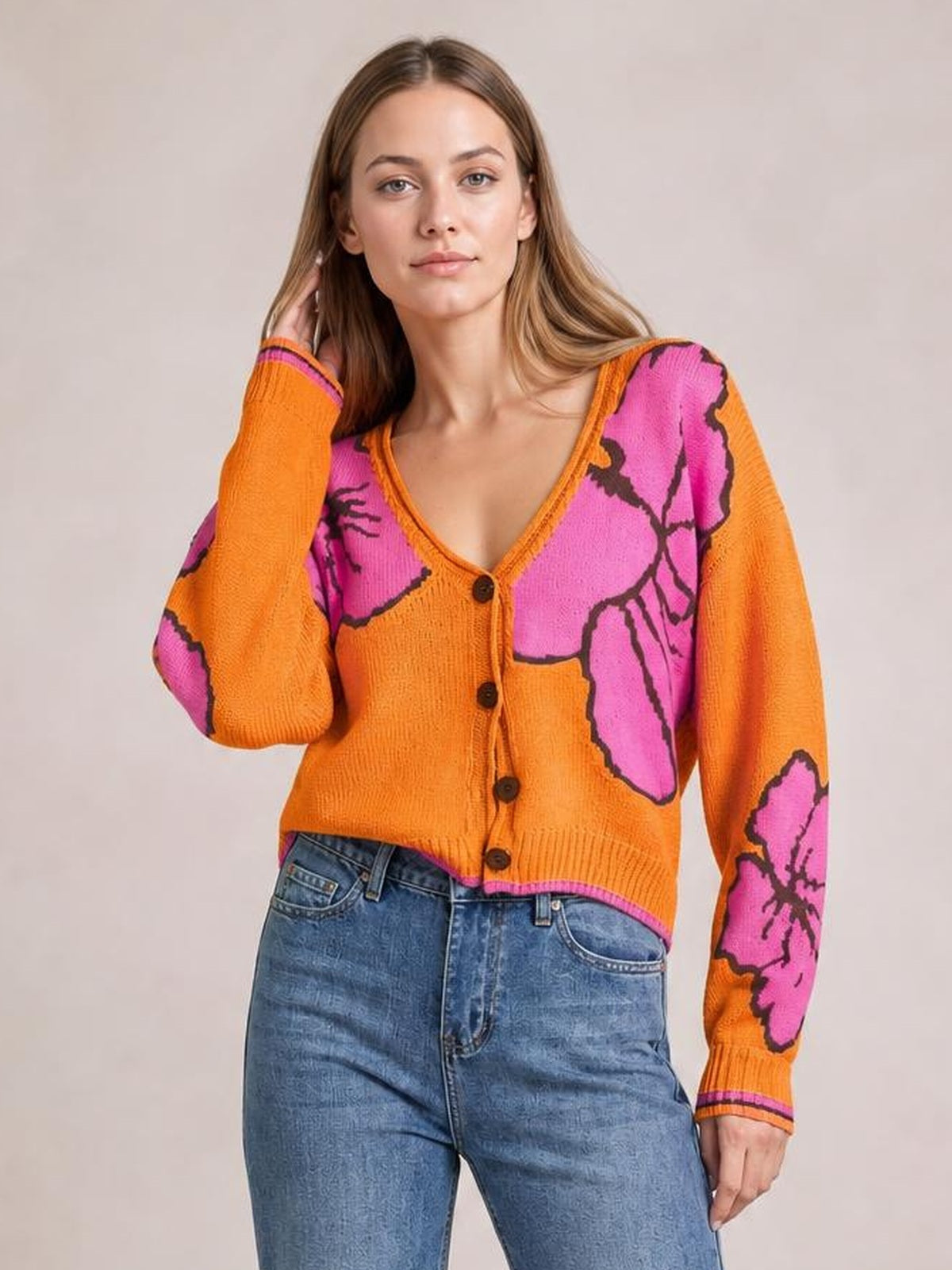 Model poses in blue jeans wearing a cropped orange knit cardigan with a V-neck, button-down front, seam details, and bold pink floral patterning.