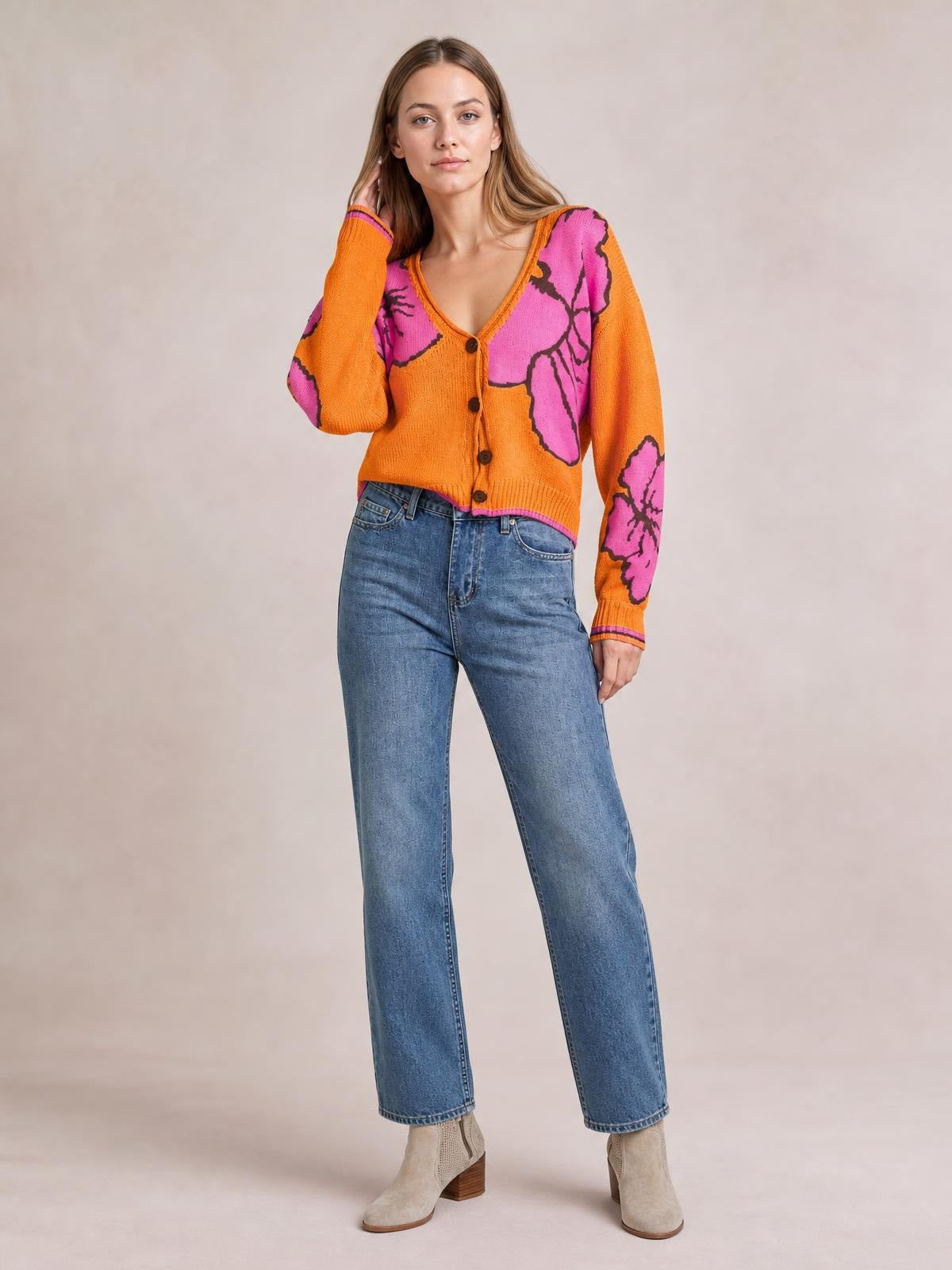 A model poses in an orange knit cardigan with a V-neck, button-down front, seam details, and bold pink and black floral motifs.