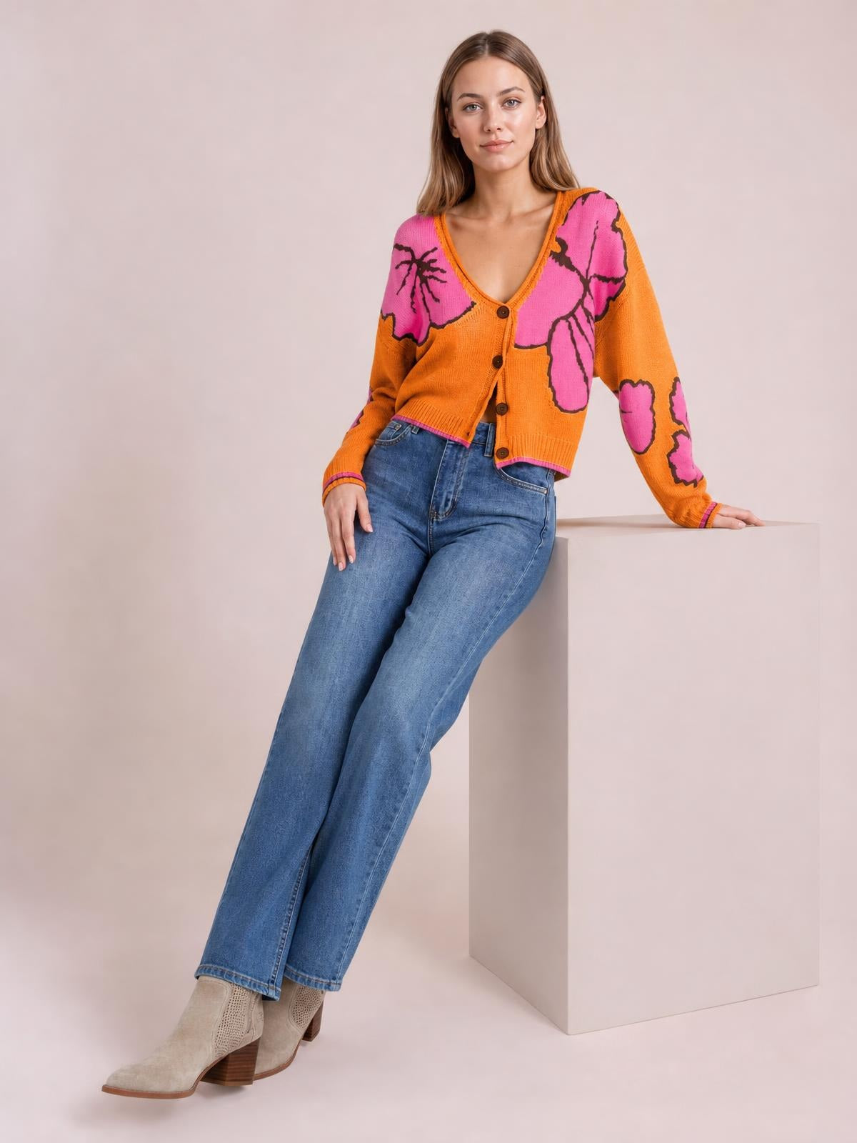 A model leans against a pedestal wearing an orange and pink floral V-neck button-down knit cardigan with long sleeves, seam details, and a cropped silhouette.