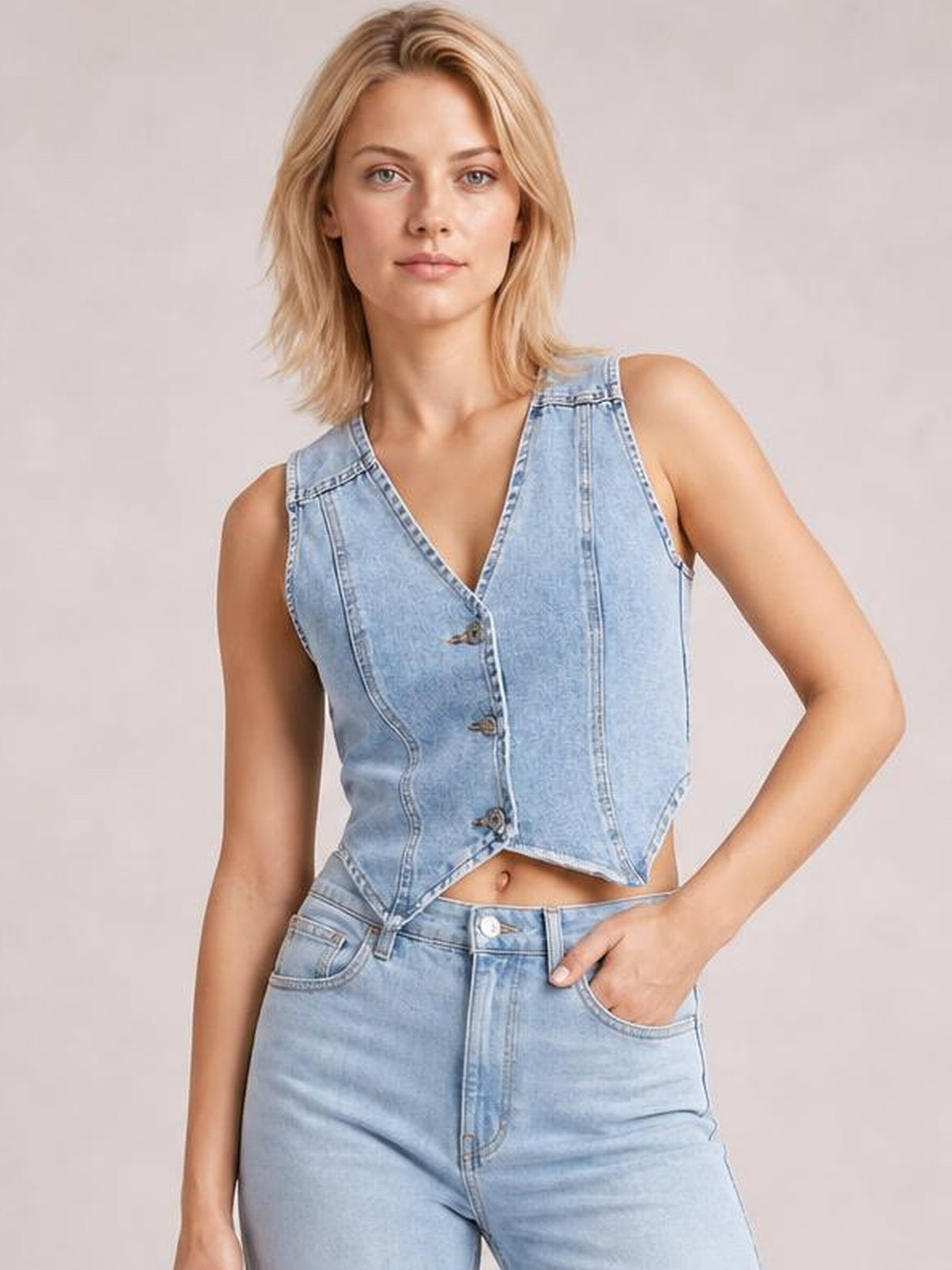 Model poses with one hand in her pocket while wearing a light blue V-neck button-up denim vest with seamed construction and a fitted cropped silhouette.