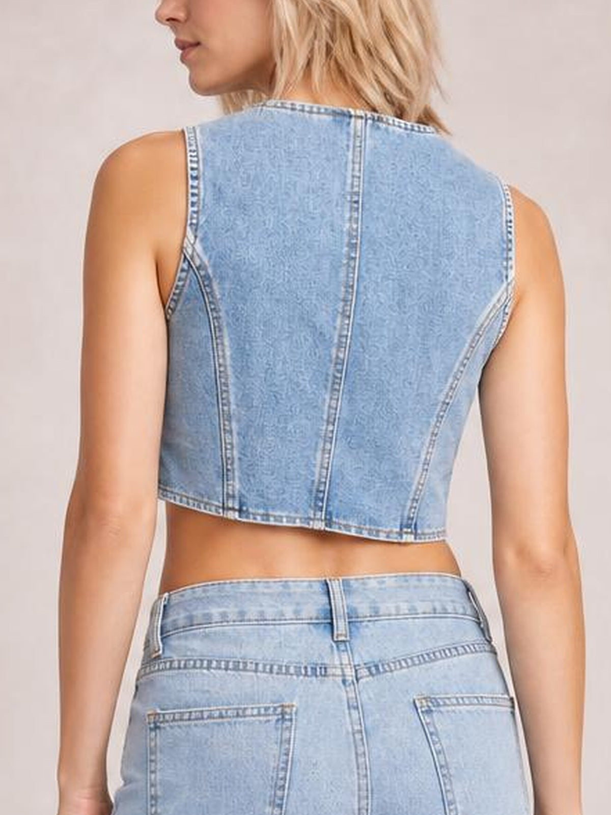 A model stands in a light blue denim vest shown from the back, featuring a sleeveless cropped silhouette and structured vertical seam construction.